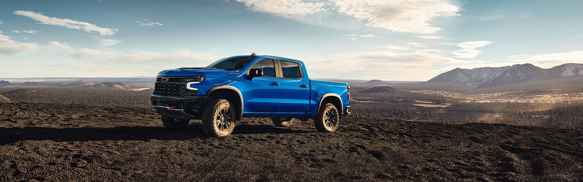 2025 Chevrolet Silverado 1500 in bright blue parked on rugged off-road terrain with mountain landscape in the background.