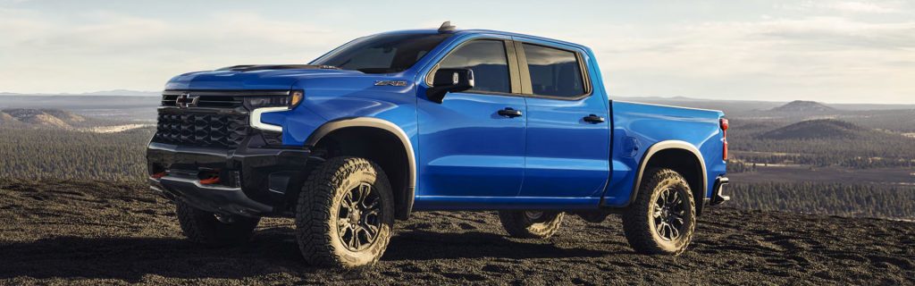 2026 Chevrolet Silverado in blue positioned on a rocky overlook, showcasing off-road capability, bold front grille, and rugged truck design.