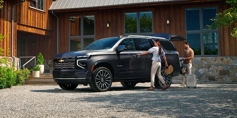 A man and woman load groceries into a black 2026 Chevrolet Tahoe parked in a driveway.