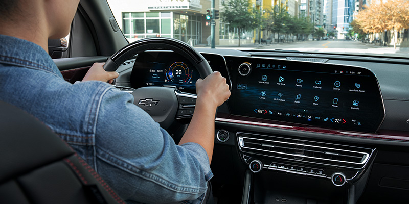 A person drives a 2026 Chevrolet Traverse with a sleek, modern dashboard featuring a large touchscreen and digital speedometer. The street outside is lightly trafficked.