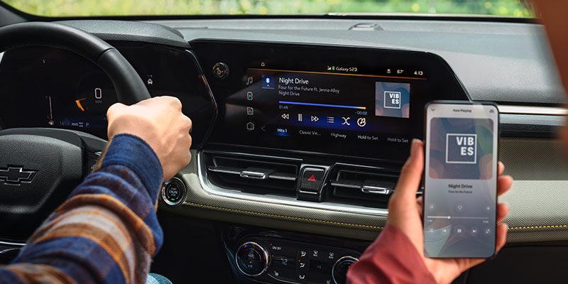 Inside a Chevrolet Trailblazer, a person drives while holding a smartphone displaying music synced to the car's touchscreen, showing "Night Drive."
